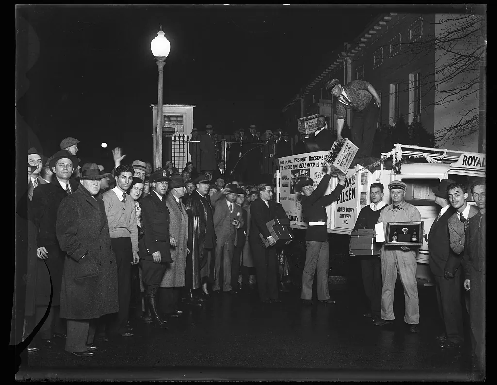 A crowd at night watches men unload cases of beer from a Royal Pilsen brewery truck