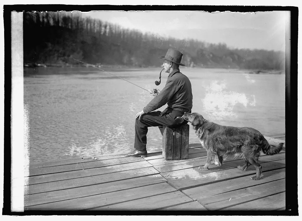 A dog sniffs the back pocket of a man fishing from a dock on the Potomac River